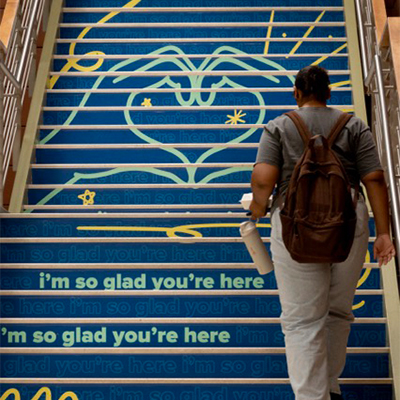 Person with backpack climbing blue stairs painted with yellow heart and repeated welcoming text