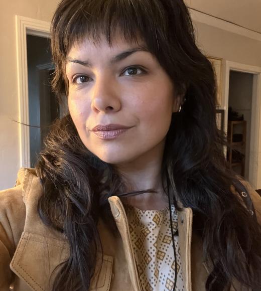 Selfie of woman with dark wavy hair and bangs, wearing tan jacket, warm indoor light