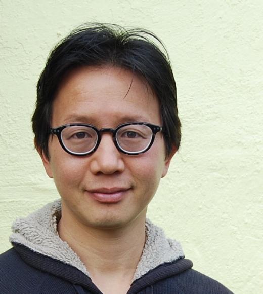 Headshot of person with round glasses, dark hair, faint smile, pale green background