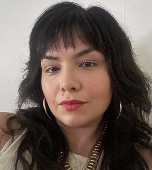 Photo of woman with gold jewelry, long brown hair and bangs, and beige top. 
