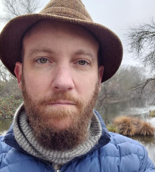Headshot: Lewis Lawyer standing by Putah Creek in a tweed hat and blue puffer jacket