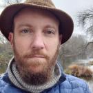 Headshot: Lewis Lawyer standing by Putah Creek in a tweed hat and blue puffer jacket